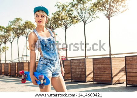 Young alternative girl skater wearing cap standing on the city street holding penny board looking aside pensive