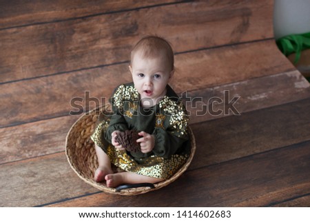 One year old girl sitting in a wicker basket. In a green dress with gold sequins and bare legs. On wooden background. Emotions. Bare foot. 