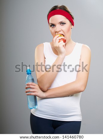 Attractive young woman in fitness wear. Woman holding a bottle of mineral water and biting red apple. Isolated on a gray background