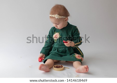 One year old girl in a green dress and bare feet sitting, eating cake. The girl was smeared with cream. Green dress with gold sequins. Dirty hands in a child. Headband. On white background. Smash cake