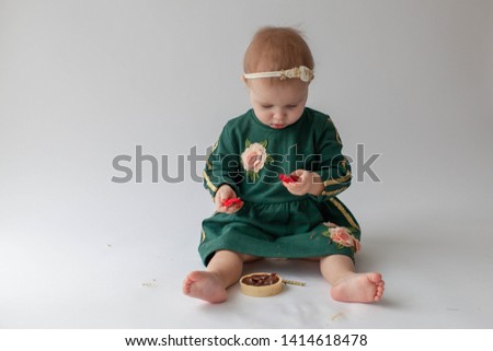 One year old girl in a green dress and bare feet sitting, eating cake. The girl was smeared with cream. Green dress with gold sequins. Dirty hands in a child. Headband. On white background. Smash cake