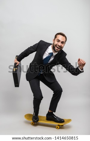 Full length of an attractive smiling young businessman wearing suit isolated over gray background, carrying briefcase while riding skateboard
