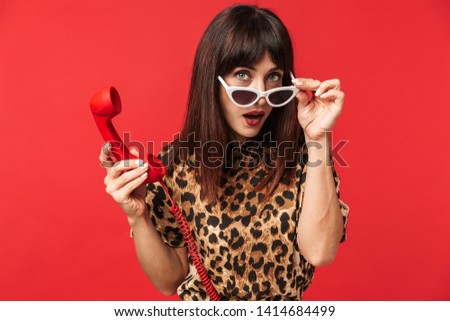 Image of a beautiful young woman dressed in animal printed shirt posing isolated over red background wearing sunglasses talking by telephone.