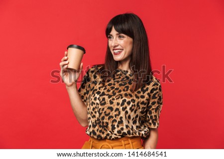 Image of a beautiful happy young woman dressed in animal printed shirt posing isolated over red background drinking coffee.