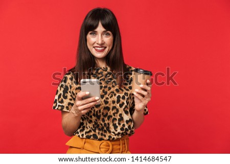 Image of a beautiful happy young woman dressed in animal printed shirt posing isolated over red background drinking coffee using mobile phone.
