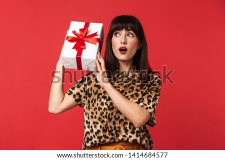 Image of a beautiful excited shocked young woman dressed in animal printed shirt posing isolated over red background holding present box.