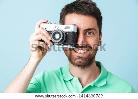 Portrait of a handsome bearded man wearing casual clothing standing isolated over blue background, taking a picture with photo camera