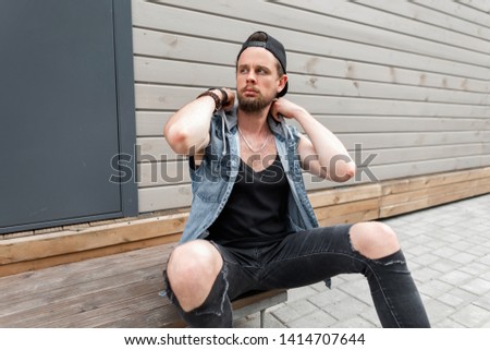Urban attractive young man with a beard in a stylish cap in a fashionable denim vest in a black t-shirt sitting near a vintage wooden wall. American guy enjoying a warm summer day. Street style.
