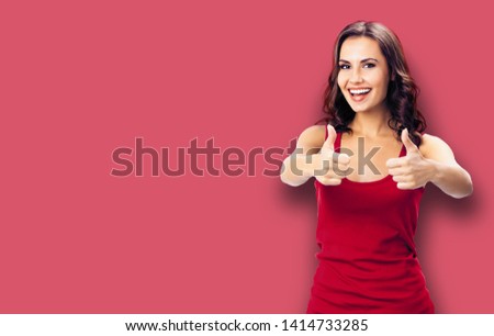 Portrait of young smiling woman in casual clothing, showing thumb up gesture, over color wall background. Happy girl in red dress. Brunette model at studio picture. Copy space area for some sign text.