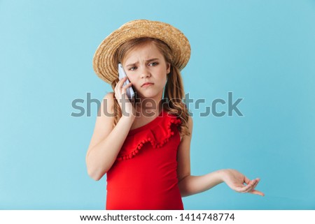 Shocked upset little girl wearing swimsuit and summer hat standing isolated over blue background, using mobile phone