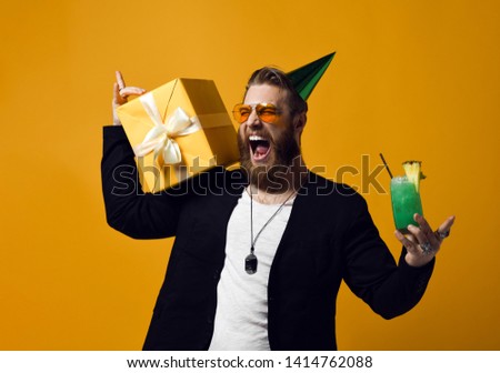 handsome bearded man with cone hat beard holding glass of cocktail and gift box celebrating birthday among friends isolated on yellow. Party