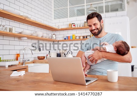Multi-Tasking Father Holds Sleeping Baby Son And Works On Laptop Computer In Kitchen Royalty-Free Stock Photo #1414770287