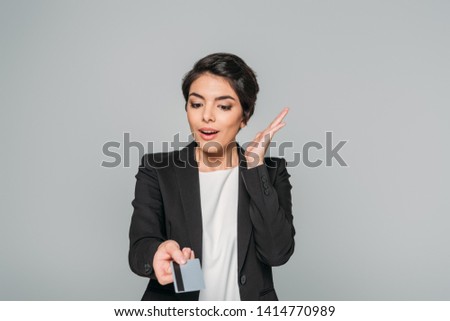 amused mixed race businesswoman gesturing while holding credit card isolated on grey