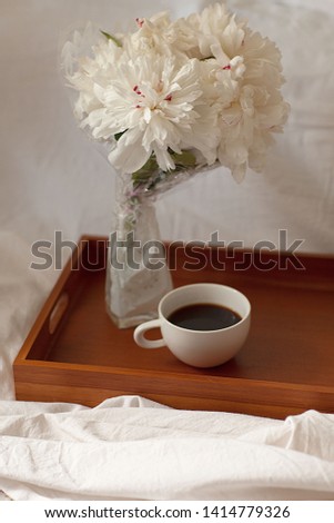 Summer morning coffee in bed. Coffee mug and a bouquet of white peonies on a wooden tray over bed linen and blanket background, selective focus . Concept of summer mood
