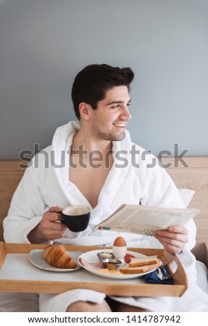 Photo of young happy man wearing white bathrobe having breakfast and reading newspaper while sitting on bed in hotel apartment