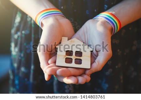 young female hands with LGBT rainbow ribbon wristbands holding craft wooden house Royalty-Free Stock Photo #1414803761