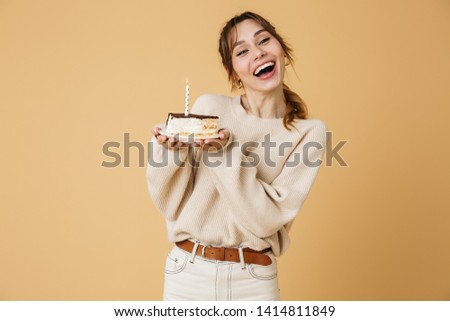 Beautiful happy young woman wearing sweater standing isolated over beige background, holding birthday cake with candle on a plate