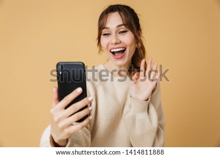 Beautiful excited young woman wearing sweater standing isolated over beige background, having a video call