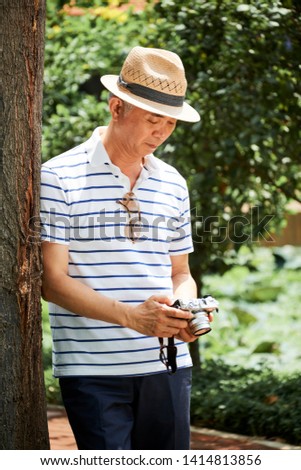 Asian senior photographer standing near the tree and watching some photos on his digital camera in summer day outdoors