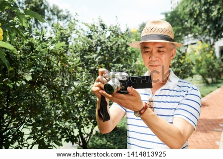 Asian senior man in hat standing in green garden and looking at display of digital camera after taken the pictures