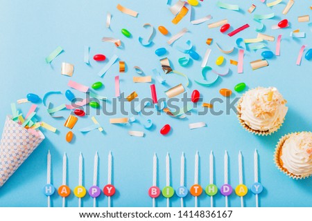 birthday hat with confetti and cupcake with burning candle on blue paper background