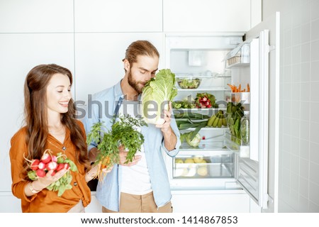 Portrait of a young and happy couple standing with fresh vegetables near the refrigerator full of healthy products at home