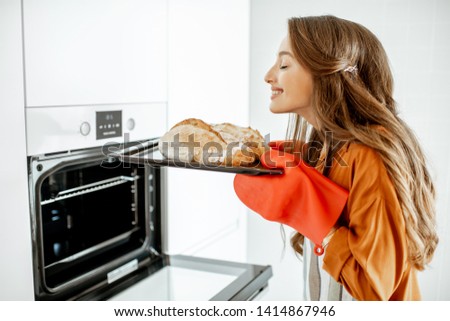 Beautiful young woman baking bread in the oven at the modern white kitchen at home Royalty-Free Stock Photo #1414867946