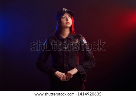Serious female police officer is posing for the camera against a black background with red and blue backlighting.