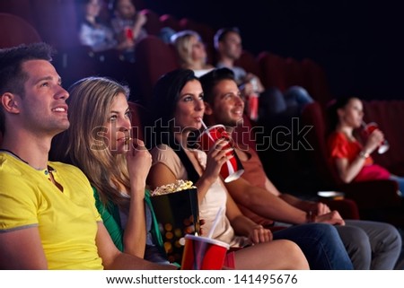 Young people sitting in multiplex movie theater, watching movie, eating popcorn.