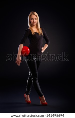 Pretty blonde dancer holding red ball, wearing black outfit over black background.