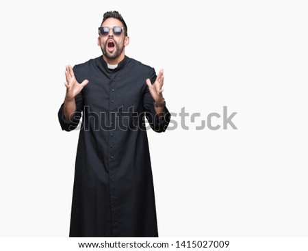 Young Christian priest wearing sunglasses over isolated background crazy and mad shouting and yelling with aggressive expression and arms raised. Frustration concept.