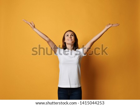 Young smiling woman holds her spread hands up with open palms and looks up like she is waiting for something fall from skies or like she is holding a textor banner above her head on yellow background 