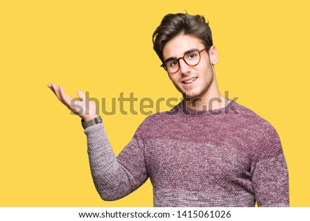 Young handsome man wearing glasses over isolated background smiling cheerful presenting and pointing with palm of hand looking at the camera.