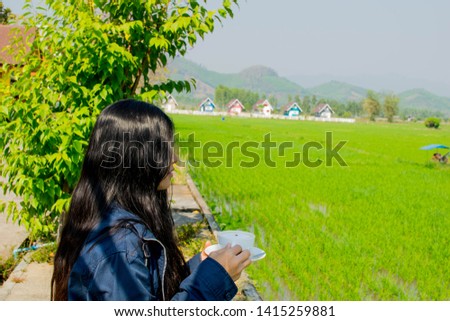 Closeup picture of Asian girl drinking coffee from a white ceramic cup with a saucer outdoor on nature with a green grass field, mountains and beautiful cute colorful houses on the horizon