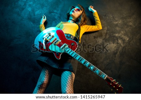 Fashion young hipster woman with curly hair with red guitar in neon lights. Rock musician is playing electrical guitar. 90s style concept.