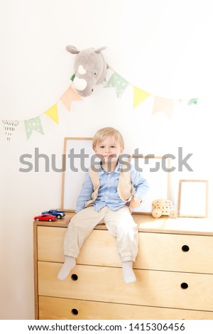 A little boy is sitting on a dresser near a white wall with flags and toys. Portrait of a boy sitting in the children's room in the Scandinavian style. Environmentally friendly children's room decor