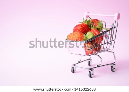Shopping trolley and red strawberry on pink background with copy space. Healthy food and summer berries concept. Sale, discount, shopaholism concept. Consumer society trend. Creative design.