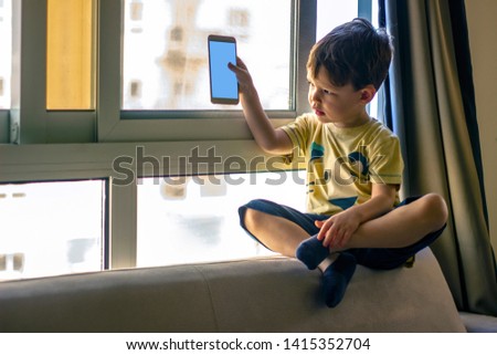 Little boy watching cartoons on mobile phone, kid with smiling face sitting on sofa and playing games on smart phone. Happy toddler boy having fun playing game on mobile phone.
