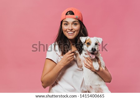 The girl in the orange baseball cap, wearing a light pink t-shirt and jeans, holds the dog by the paw sitting on her lap, smiling widely with surprised open eyes over pink wall. Happiness and dreams. Royalty-Free Stock Photo #1415400665