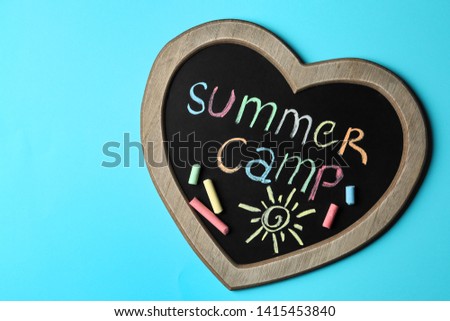 Heart shaped blackboard with text SUMMER CAMP, drawing and chalk sticks on color background, top view. Space for design