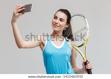 Smiling woman tennis player holding racket isolated over gray background, taking selfie