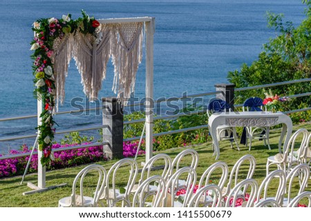 Wedding arch in Boho style near the ocean decorated with macrame and fresh flowers, chairs, signing table and petal aisle  for a ceremony. Concept of a tropical wedding. Bali, Indonesia.
