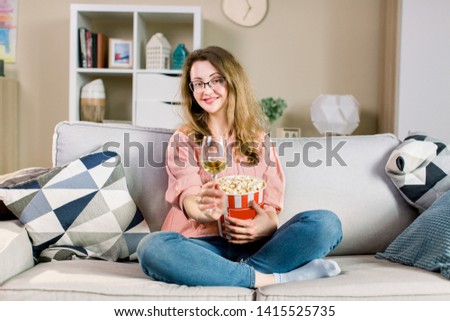A beautiful young blond woman sitting on a couch, drinking wine and eating popcorn, smiling