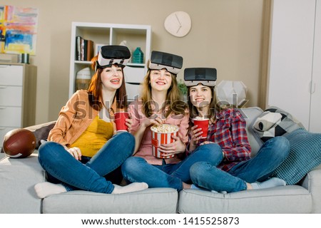 Three young attractive woman wearing headset VR virtual reality vision goggles, having party at home, eating popcorn