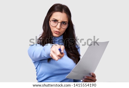 Portrait of brunette woman wearing blue long sleeve blouse and transparent eyewear, writing on white blank paper with copy space for your advertisement information, against white studio wall.