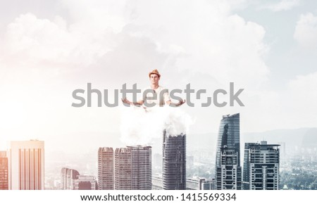 Man in white clothing keeping eyes closed and looking concentrated while meditating on cloud in the air with cityscape view on background.