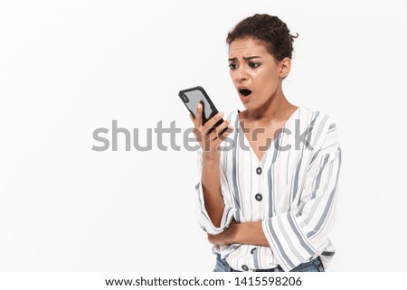 Attractive shocked young african woman wearing casual clothes standing isolated over white background, holding mobile phone