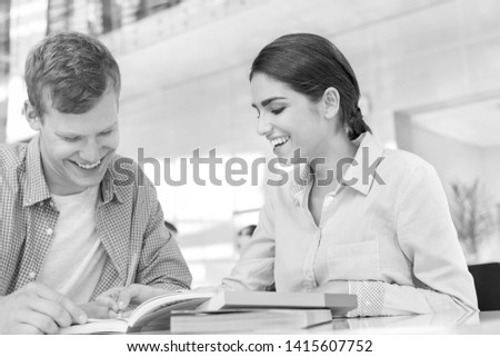 Black and White photo of Smiling friends discussing over book while sitting at table in library