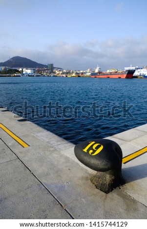Big mooring bollard in a commercial harbour, port basin water and ships in background.