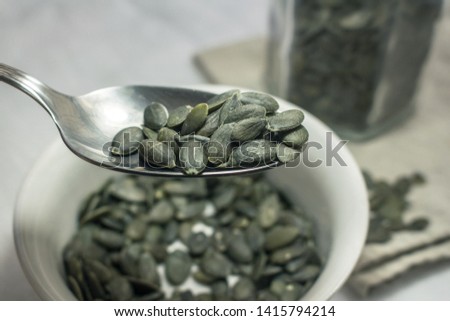detail of a spoon full of pumpkin seeds, in the background a bowl, a glass jar and other seeds scattered on a light cotton napkin
